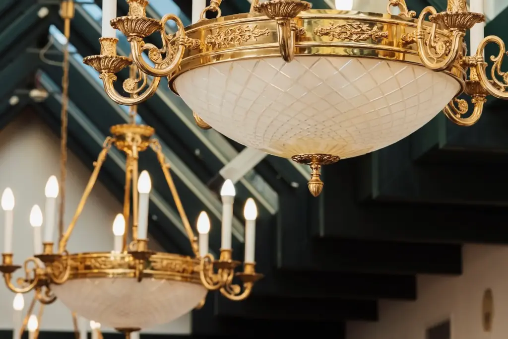 Zwei verschnörkelte goldene Kronleuchter mit weißen Glasschirmen und mehreren Glühbirnen hängen von der Decke, mit dunklen Balken und natürlichem Licht im Hintergrund.