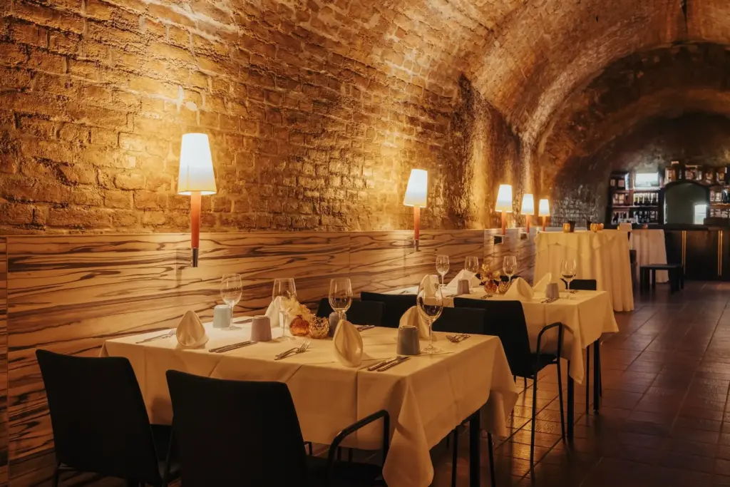 Ein gemütliches Restaurant mit gewölbten Ziegeldecken, sanft beleuchtet von Wandlampen. Die Tische sind mit weißen Tüchern bedeckt und mit Gläsern, Tellern und Servietten gedeckt, was eine einladende, elegante Atmosphäre schafft.