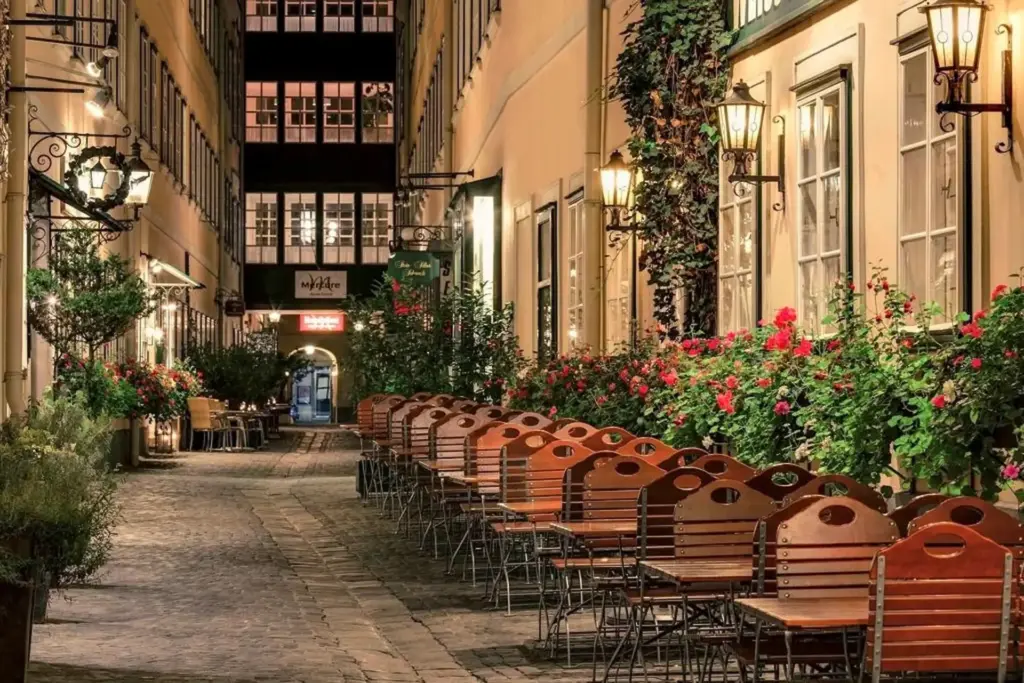 Eine charmante Gasse mit Kopfsteinpflaster, gesäumt von leeren Holztischen und -stühlen vor einem Café, gesäumt von leuchtend roten Blumen und nachts warm beleuchtet von klassischen Wandlaternen.