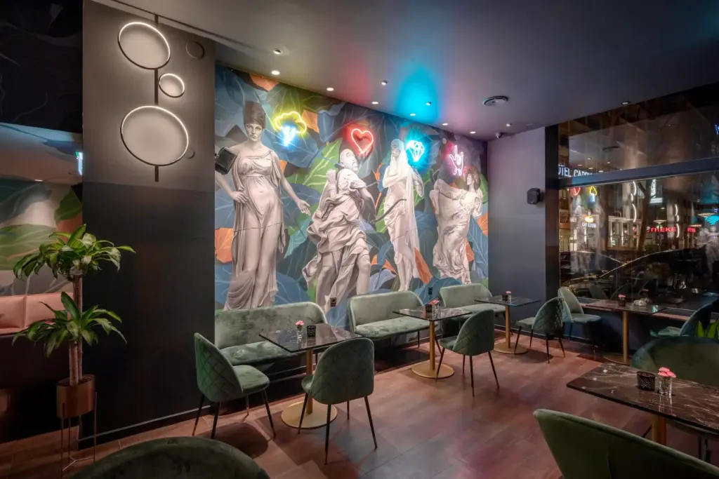 Ein modernes Café mit samtgrünen Stühlen, kleinen Tischen und einem farbenfrohen Wandgemälde mit klassischen Statuen an der Wand, beleuchtet von Neonröhren in Form einer Krone, eines Herzens und eines Blitzes.