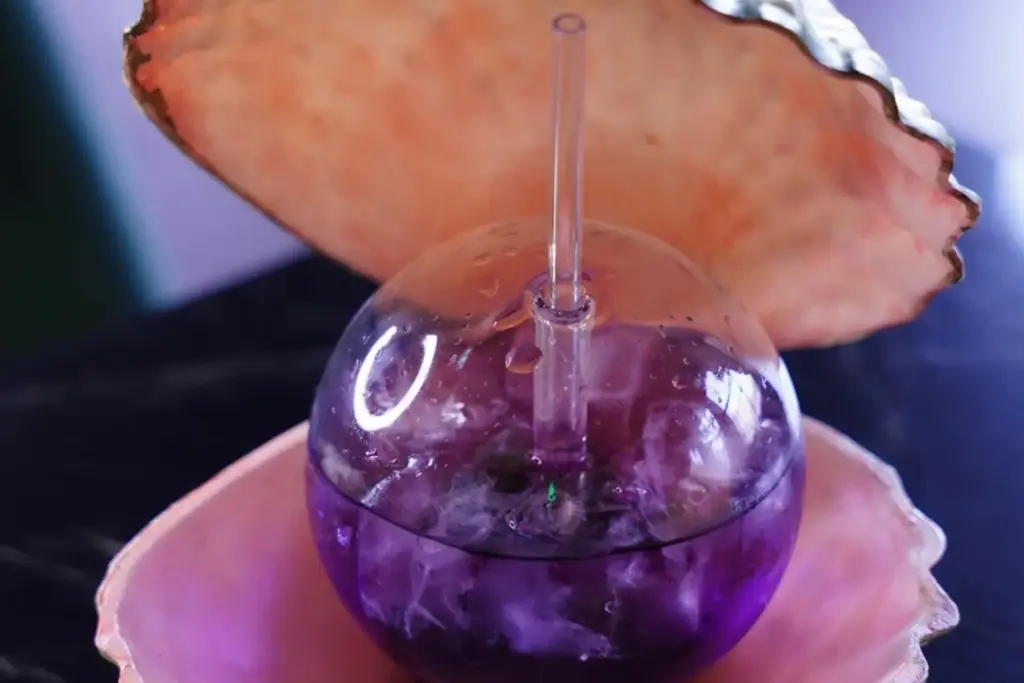 Ein rundes Glas, gefüllt mit einem lilafarbenen eisgekühlten Getränk, steht in einer rosafarbenen muschelförmigen Schale, mit einem durchsichtigen Strohhalm und einem offenen Muscheldeckel im Hintergrund.