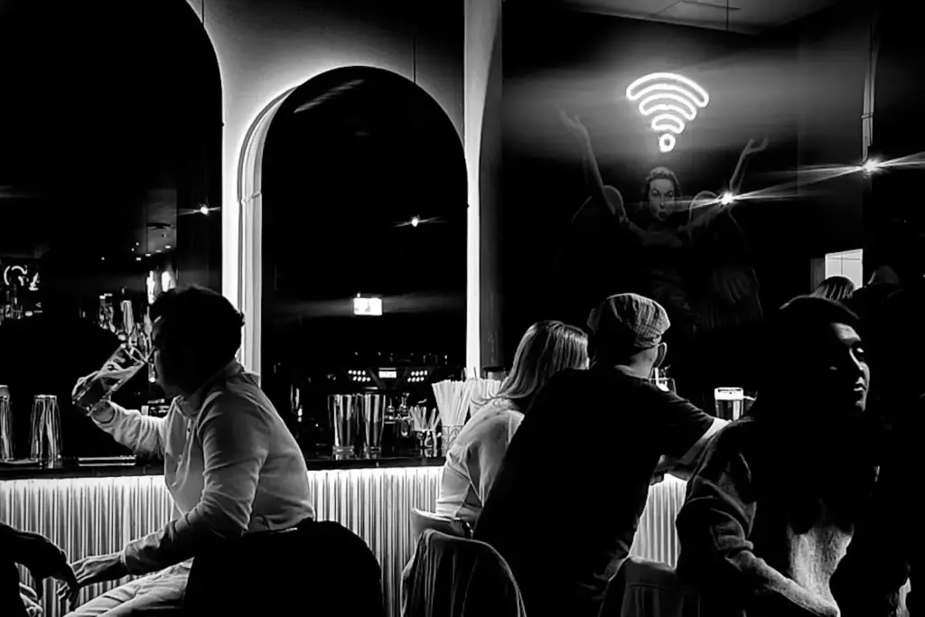 Schwarz-Weiß-Foto von Menschen, die an einer Bar sitzen, einige reden, andere schauen weg. Ein neonfarbenes Wi-Fi-Symbol leuchtet über einem Wandgemälde, das eine Person mit erhobenen Armen zeigt, an der Wand im Hintergrund.