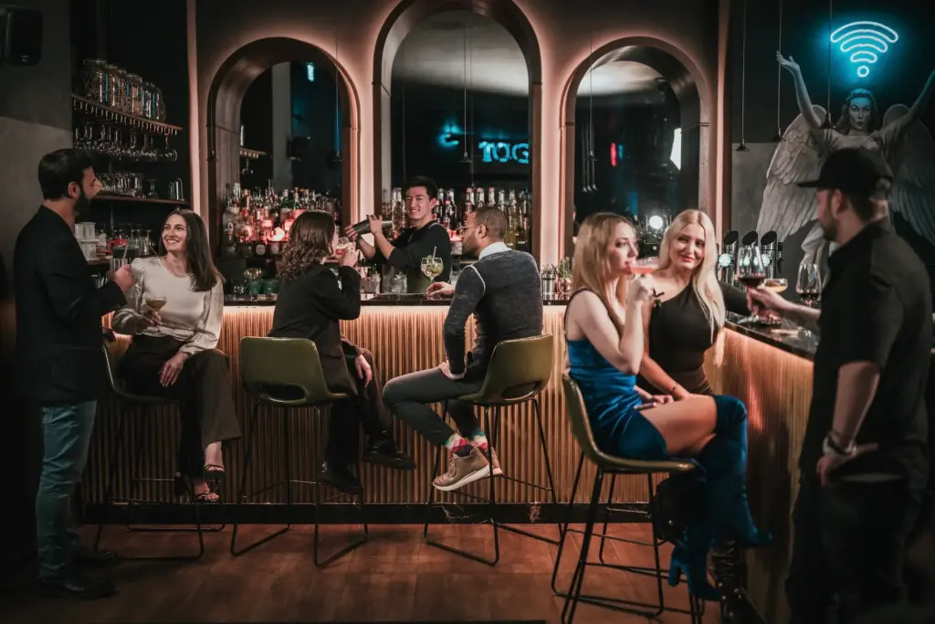 In einer stilvollen Bar mit schummriger Beleuchtung kommen die Menschen zusammen. Einige sitzen auf Barhockern, andere stehen, plaudern und trinken. An der Wand hinter dem Tresen leuchtet ein Neonschild im Toga Wien.