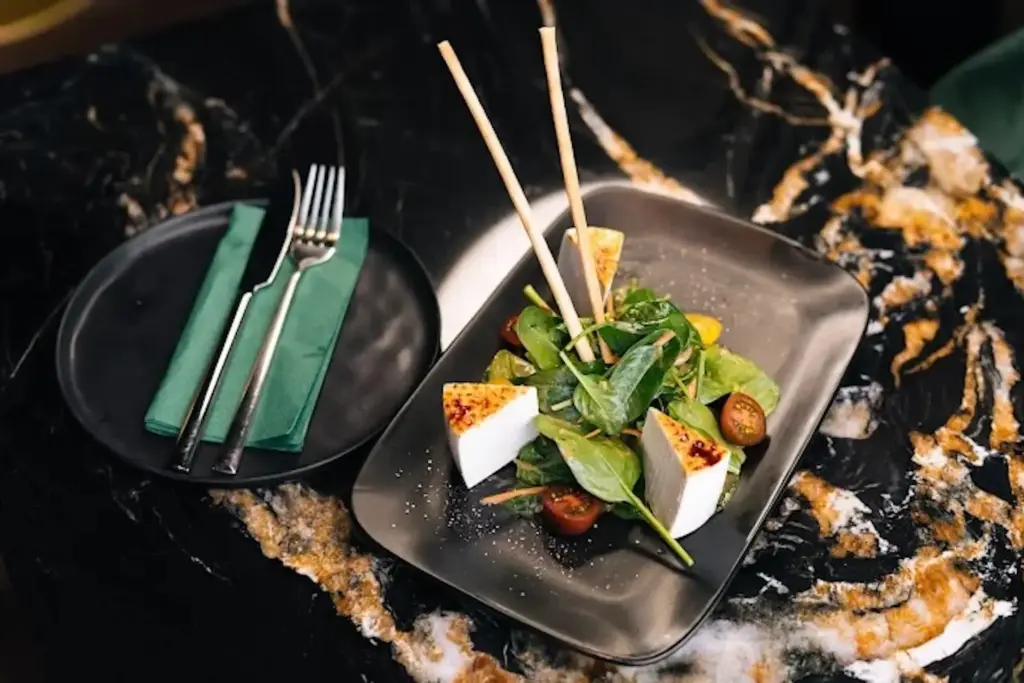 Ein modern gedeckter Tisch mit einem schwarzen Salatteller mit frischem Spinat, Kirschtomaten, gegrillten Käsedreiecken und zwei Stäbchen, daneben ein kleiner Teller mit Gabel und Messer, eingewickelt in eine grüne Serviette.
