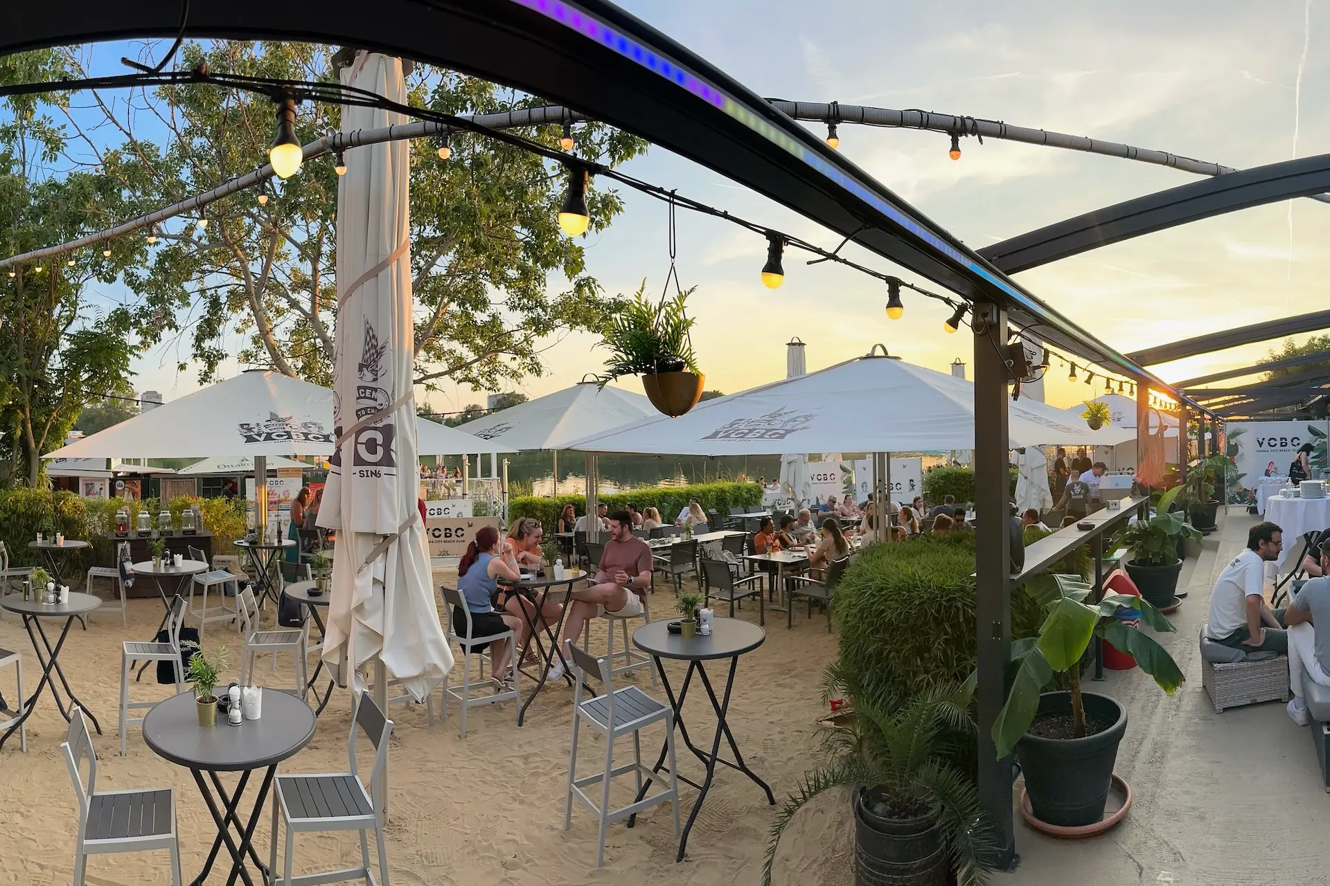 Im Vienna City Beach Club sitzen die Leute an Tischen im Freien auf einer Sandterrasse unter großen weißen Sonnenschirmen und entspannen sich. Lichterketten und Pflanzen schmücken den Bereich, während die Sonne im Hintergrund untergeht und eine perfekte Strandcafé-Atmosphäre schafft.