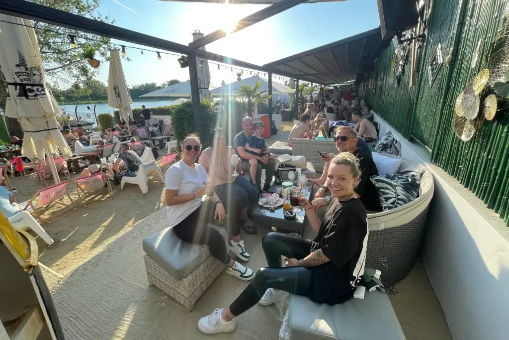 Eine Gruppe von vier Personen lächelt in einer Outdoor-Lounge am Strand und genießt Getränke und Snacks in der Sonne. Andere entspannen sich an Tischen in der Nähe. Sonnenschirme und Grünpflanzen sorgen für eine lebhafte, entspannte Atmosphäre.